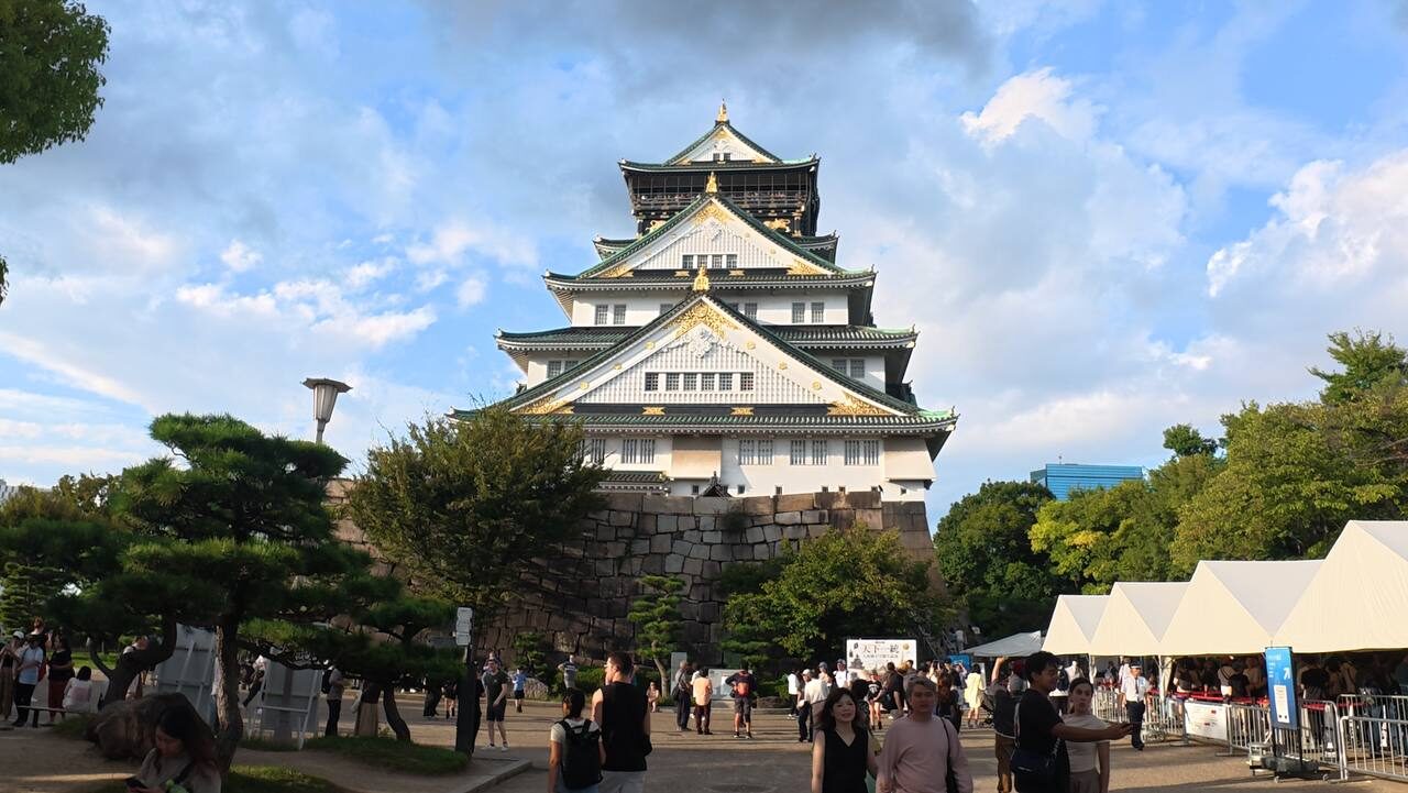 Osaka Castle main keep, white walls and gold accents under a blue sky with scattered clouds, pine trees in the foreground.